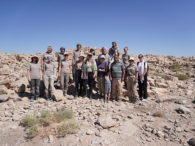 Das Bild zeigt eine Gruppe von 17 Personen und einem kleinen Kind, vor dem "Tel" (Hügel) der antiken Siedlung al-Jumayil. Bei den Personen handelt es sich um die Teilnehmer_Innen der Lehrgrabung, sowie die österreichische Botschafterin und eine ihrer Mi